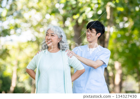 Glasses asian man caregiver nurse shoulder massaging old woman granny patient while standing in park Glasses asian man caregiver nurse shoulder massaging old woman granny patient while standing in park 134327430