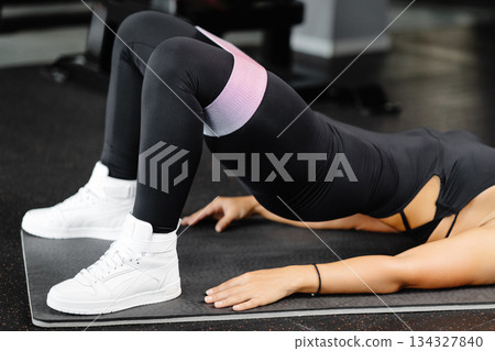 Young woman in black sportswear training with elastic resistance band above knees in gym. Lady doing glute bridge exercise for buttocks, legs and core 134327840