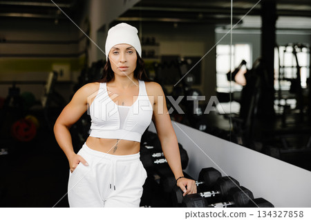 Beautiful fit woman looking at camera after gym exercise. Middle aged woman wearing sports bra and pants standing near simulator in gym Beautiful fit woman looking at camera after gym exercise. Middle aged woman wearing sports bra and pants standing near simulator in gym 134327858