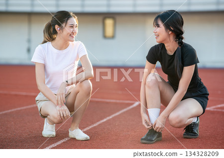 Pretty asian woman crouching talking to friend tying sneaker lace on running track in sports stadium 134328209