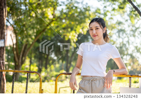 Pretty asian woman smiling while standing leaning against a yellow rusty fence in the park or garden 134328248