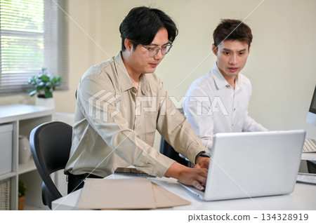 Glasses asian businessman typing or showing laptop screen to coworker at table in company's office. 134328919