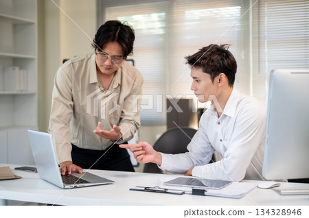 Glasses asian businessman talking to coworker while standing aside laptop at table in company office Glasses asian businessman talking to coworker while standing aside laptop at table in company office 134328946