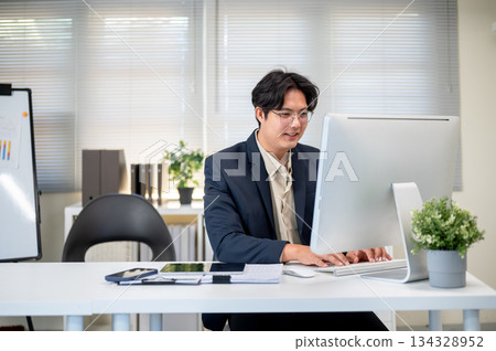 Smiling glasses asian businessman typing or looking at computer on working table in company's office 134328952