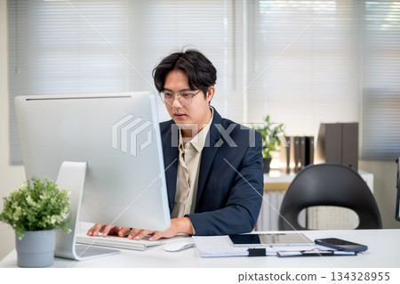 Serious glasses asian businessman typing or looking at computer on working table in company's office Serious glasses asian businessman typing or looking at computer on working table in company's office 134328955