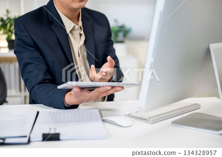 Close up of businessman holding tablet and looking at computer on working table in company's office. Close up of businessman holding tablet and looking at computer on working table in company's office. 134328957
