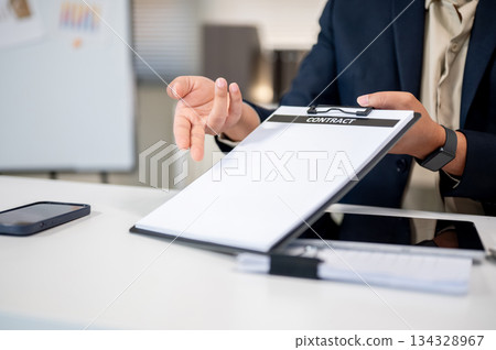 Close up of businessman's hand showing a contract document on clipboard at table in company's office Close up of businessman's hand showing a contract document on clipboard at table in company's office 134328967