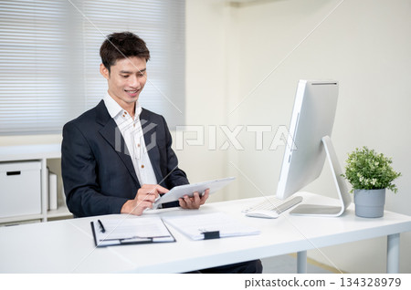 Smiling asian businessman holding and looking at tablet aside computer on table in company's office. Smiling asian businessman holding and looking at tablet aside computer on table in company's office. 134328979