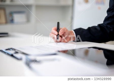 Close up businessman's hand holding pen writing in document on clipboard at working table in office. 134328990