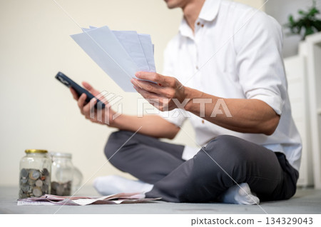 Close up of office worker man holding phone and bills as sitting on living room floor with coin jars 134329043