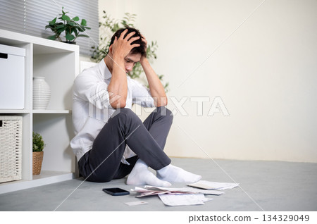 Frustrated asian man office worker holding head looking at bills as sitting on the living room floor Frustrated asian man office worker holding head looking at bills as sitting on the living room floor 134329049
