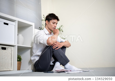 Asian man office worker holding chin thinking as looking at bills while sitting on living room floor 134329050