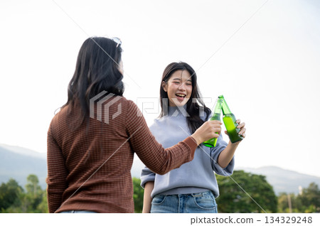 Asian woman and friend standing holding drink clinking bottles as go on picnic with mountain and sky Asian woman and friend standing holding drink clinking bottles as go on picnic with mountain and sky 134329248