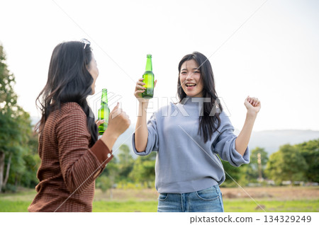 Asian woman and friend holding drink bottle dancing in green grass field with mountain view and sky. 134329249