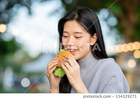 Pretty asian woman holding and eating a burger while sitting at table in a campground restaurant. Pretty asian woman holding and eating a burger while sitting at table in a campground restaurant. 134329271