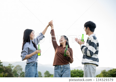 Asian woman friends holding hands dancing and man standing holding drink bottle in mount grass field Asian woman friends holding hands dancing and man standing holding drink bottle in mount grass field 134329379
