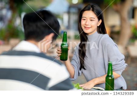 Asian woman holding alcohol drink bottle sitting across man friend at table in campground restaurant Asian woman holding alcohol drink bottle sitting across man friend at table in campground restaurant 134329389