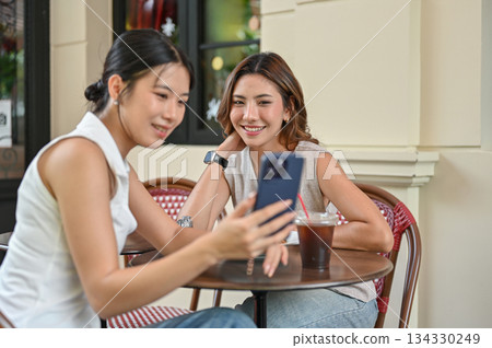 Asian woman showing pics on phone to a friend sitting at wooden table in front of cafe or restaurant 134330249