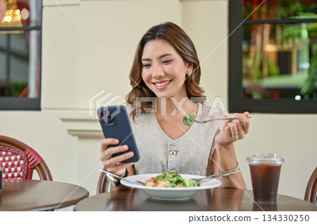 Asian woman holding fork eating salad and looking at phone sitting at table outside cafe restaurant 134330250