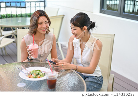 Pretty asian woman holding drink cup looking at friend's phone sits at table in a cafe or restaurant Pretty asian woman holding drink cup looking at friend's phone sits at table in a cafe or restaurant 134330252