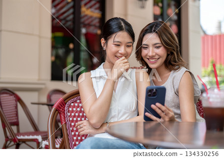Pretty asian woman showing photos on phone to a laughing friend while sitting at table outside cafe Pretty asian woman showing photos on phone to a laughing friend while sitting at table outside cafe 134330256