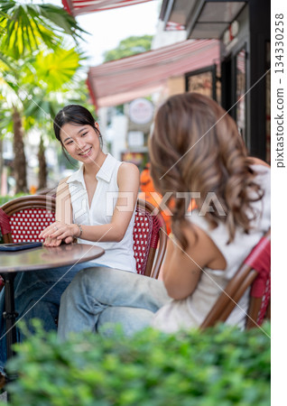Woman holding camera taking pictures of pretty asian friend while sitting at table in cafe's garden. Woman holding camera taking pictures of pretty asian friend while sitting at table in cafe's garden. 134330258