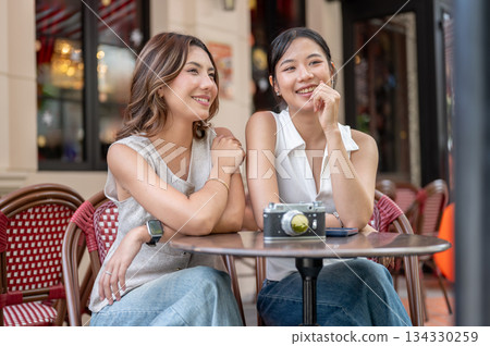 Pretty asian woman talking beside a friend while sitting with camera at round table outside the cafe Pretty asian woman talking beside a friend while sitting with camera at round table outside the cafe 134330259