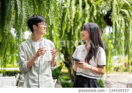 Pretty asian woman holding phone talking to a glasses man while standing under tree in cafe's garden Pretty asian woman holding phone talking to a glasses man while standing under tree in cafe's garden 134330357
