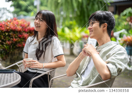 Pretty asian woman holding coffee cup sitting with glasses man friend at round table in cafe garden. Pretty asian woman holding coffee cup sitting with glasses man friend at round table in cafe garden. 134330358