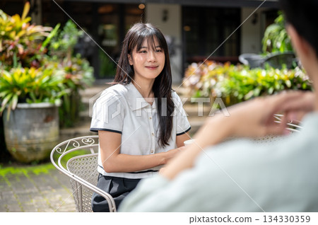 Pretty asian woman listening to man friend talking with smile while sitting at table in cafe garden. Pretty asian woman listening to man friend talking with smile while sitting at table in cafe garden. 134330359