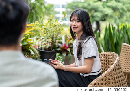 Pretty asian woman holding phone looking at man friend while sitting on woven chair in cafe's garden Pretty asian woman holding phone looking at man friend while sitting on woven chair in cafe's garden 134330362
