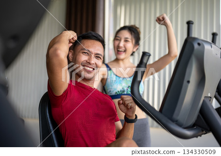 Fitness trainer coach man raising fists with a woman client while sitting on exercise equipment in gym. 134330509