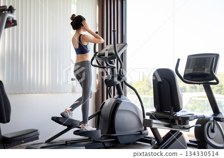 Full body back view shot of a healthy woman wearing headphones while using exercise equipment in gym 134330514