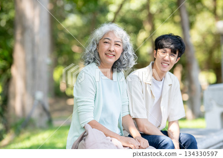 Old asian woman grandmother smiling while sitting on bench with a young man grandson in the park. 134330597