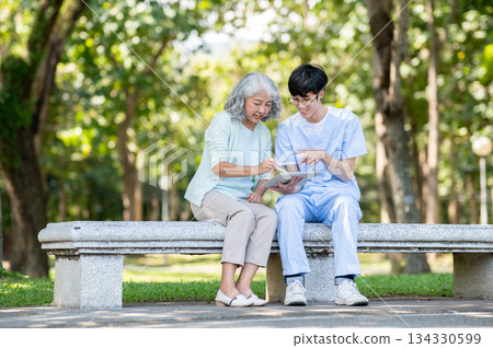 Old woman grandmother writing in tablet of young man caregiver nurse while sitting on bench in park. Old woman grandmother writing in tablet of young man caregiver nurse while sitting on bench in park. 134330599