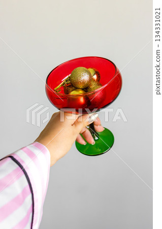 Female hand holding red glass filled with golden Christmas ornaments. Elegant festive still life for New Year, Christmas, holiday decor and celebration concepts 134331021