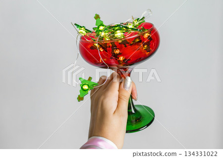Female hand holding red and green glass with glowing Christmas lights. Festive minimal composition with copy space for holiday, party or celebration concepts. 134331022