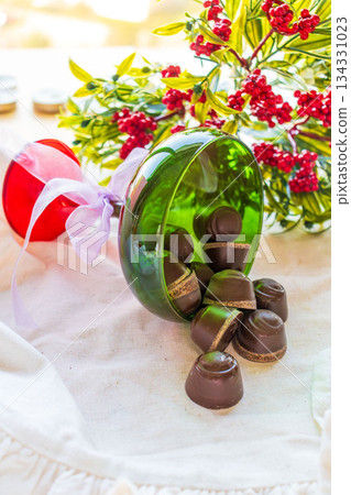 Chocolates are poured out of a green glass glass onto a white cloth with festive decor in the background. 134331023