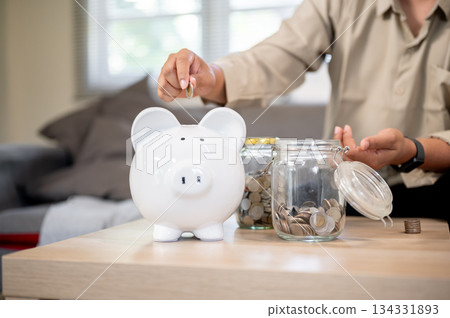 Close up of man's hand putting coin into piggy bank aside glass jars sitting on sofa in living room Close up of man's hand putting coin into piggy bank aside glass jars sitting on sofa in living room 134331893