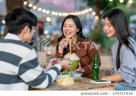 Asian woman holding coconut laughing aside friends sitting eating at table in campground restaurant. 134331910