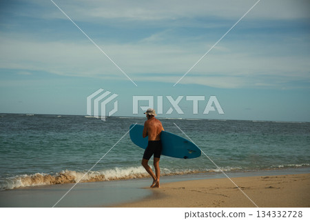 Man strolls on the beach holding a surfboard near the waters edge Man strolls on the beach holding a surfboard near the waters edge 134332728