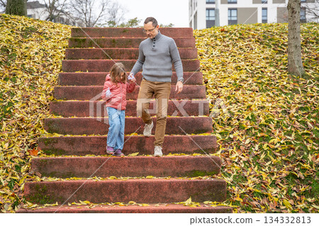 Father and daughter walking down autumn stairs in a park. Seasonal bonding and peaceful movement outdoors. Father and daughter walking down autumn stairs in a park. Seasonal bonding and peaceful movement outdoors. 134332813