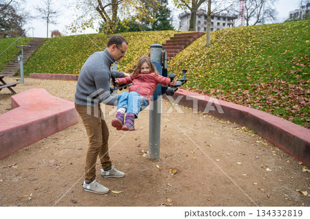 Father helping daughter to play on a playground rotating machine. Outdoor physical activity and family bonding. 134332819