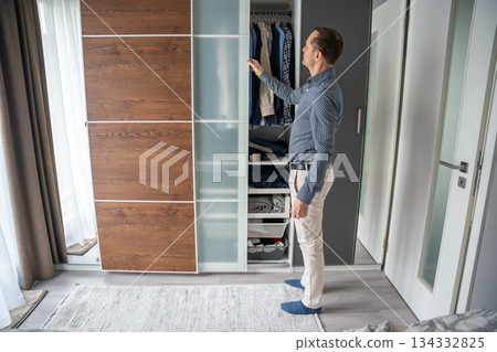 Man looking into an open closet while choosing clothes. Organizing and selecting outfit in the morning. Man looking into an open closet while choosing clothes. Organizing and selecting outfit in the morning. 134332825