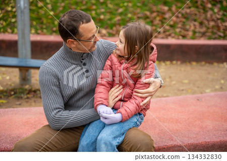 Father and daughter sitting close and talking on the playground border. Tender moment of connection and support. Father and daughter sitting close and talking on the playground border. Tender moment of connection and support. 134332830