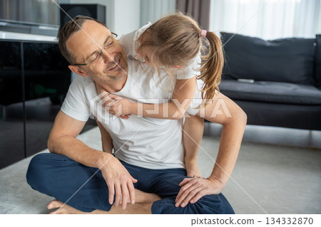 Father sitting on the carpet with daughter on his back and smiling. Everyday family interaction and parent-child affection. Father sitting on the carpet with daughter on his back and smiling. Everyday family interaction and parent-child affection. 134332870