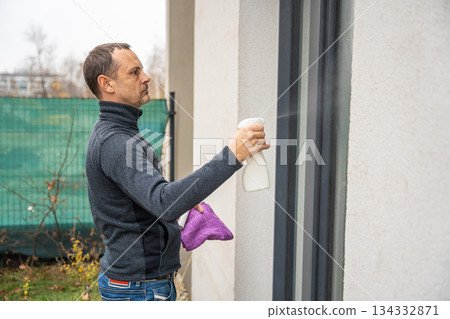 Man sprays cleaning liquid on outdoor window. Theme of hygiene and seasonal home maintenance. Man sprays cleaning liquid on outdoor window. Theme of hygiene and seasonal home maintenance. 134332871