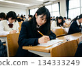 A female student with a serious expression taking a written exam in a university lecture hall and exam hall, along with other test-takers [Generative AI] 134333249