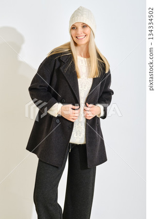 Woman dressed in warm winter coat and hat poses in casual style at indoor location during daylight Woman dressed in warm winter coat and hat poses in casual style at indoor location during daylight 134334502