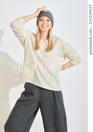 Smiling woman wearing cozy sweater and beanie poses confidently against a plain background 134334527